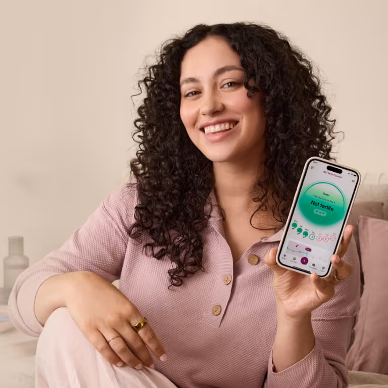 Smiling woman with curly hair holding a smartphone displaying a fertility tracking app indicating 'Not fertile'.