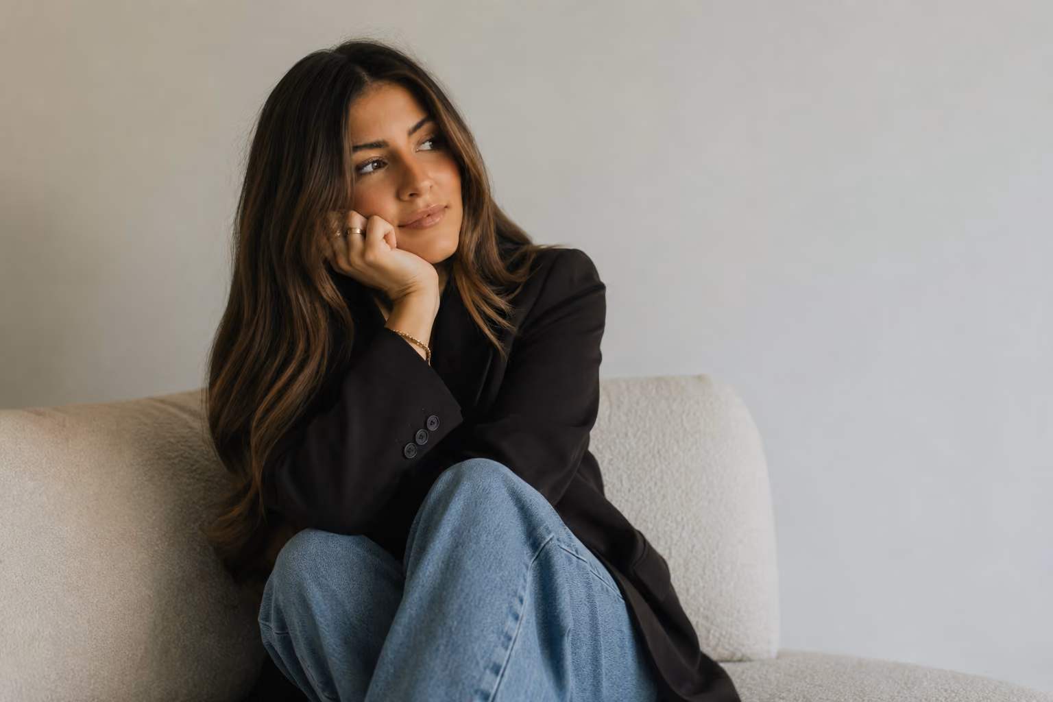 Woman with long brown hair in black blazer and jeans sitting on a couch looking thoughtfully to the side.
