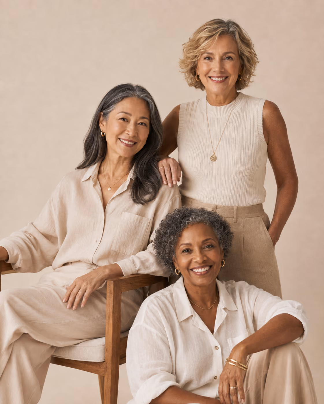 Three smiling mature women in beige and white outfits posing against a beige background.
