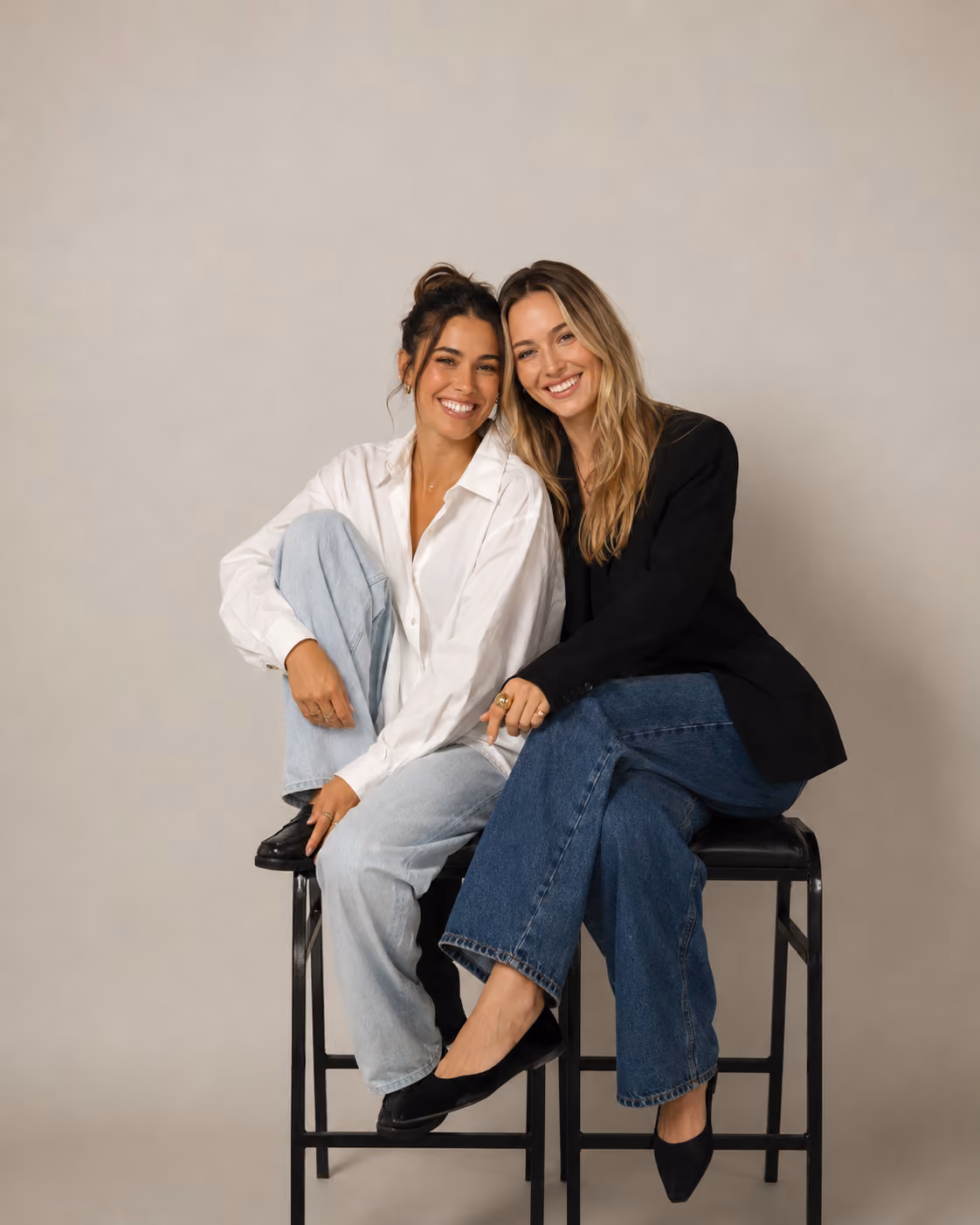Two women sitting on stools smiling, one in white shirt with light jeans, the other in black blazer with dark jeans.