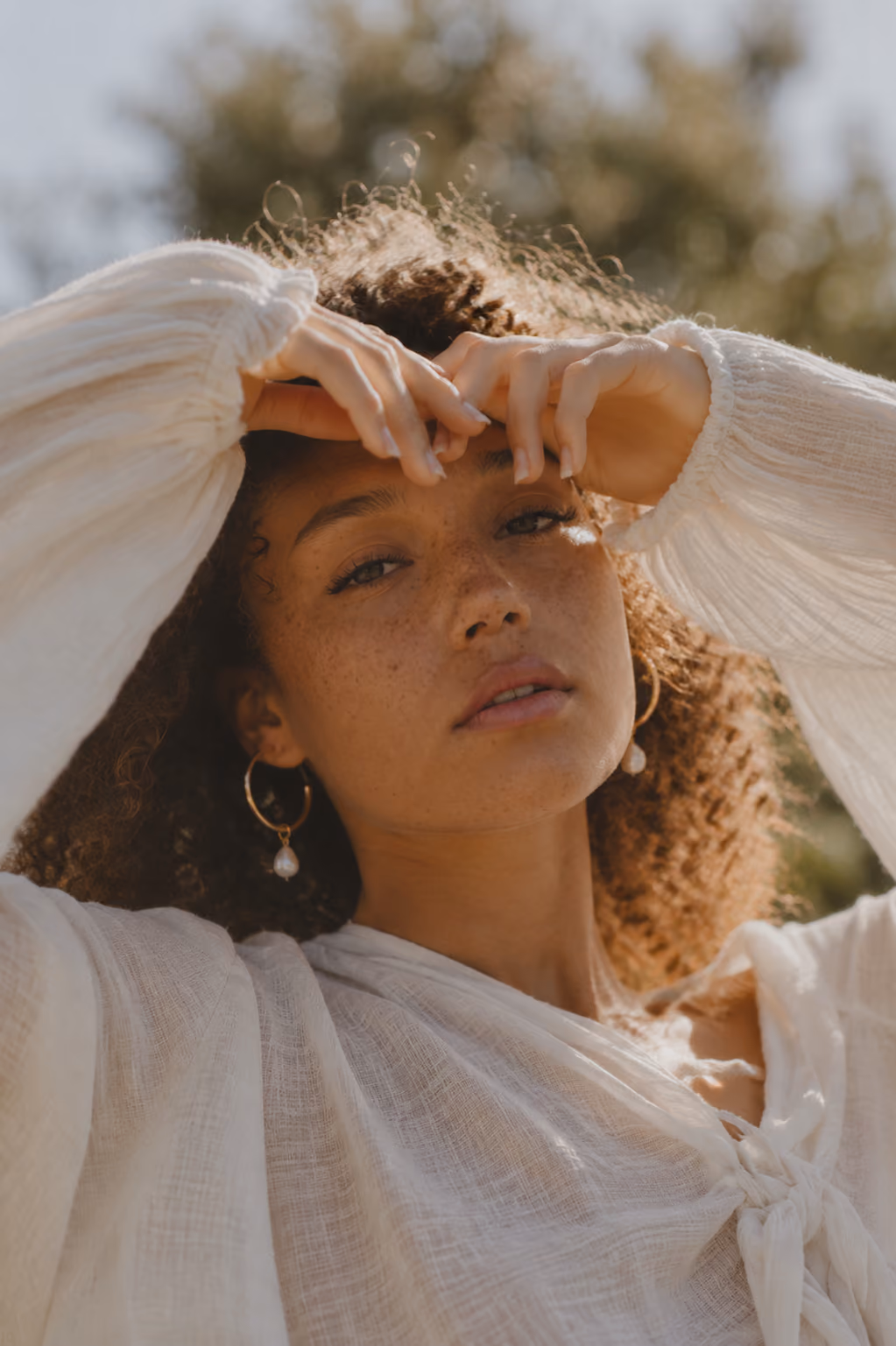 Portrait of a curly-haired woman in a white blouse posing outdoors with hands near her forehead.