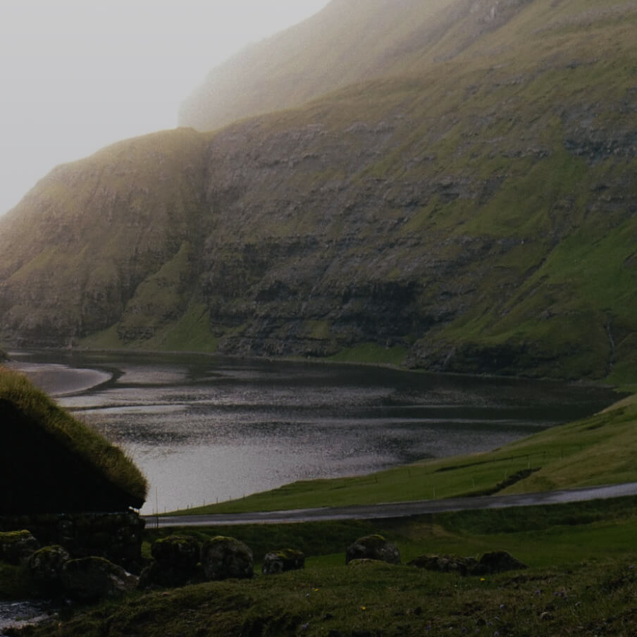 Misty lakeside scene with steep, grassy hills and a small stone building with a grass roof.