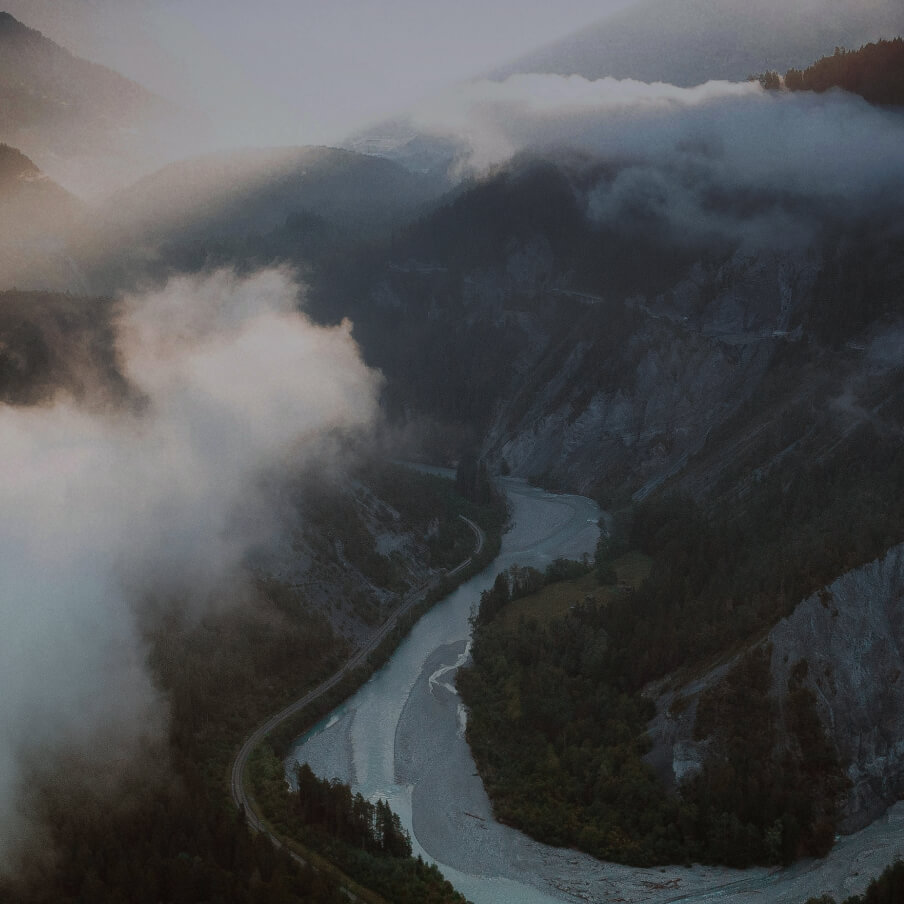 A winding river flowing through a forested valley with mist and low clouds partially covering the surrounding mountains.