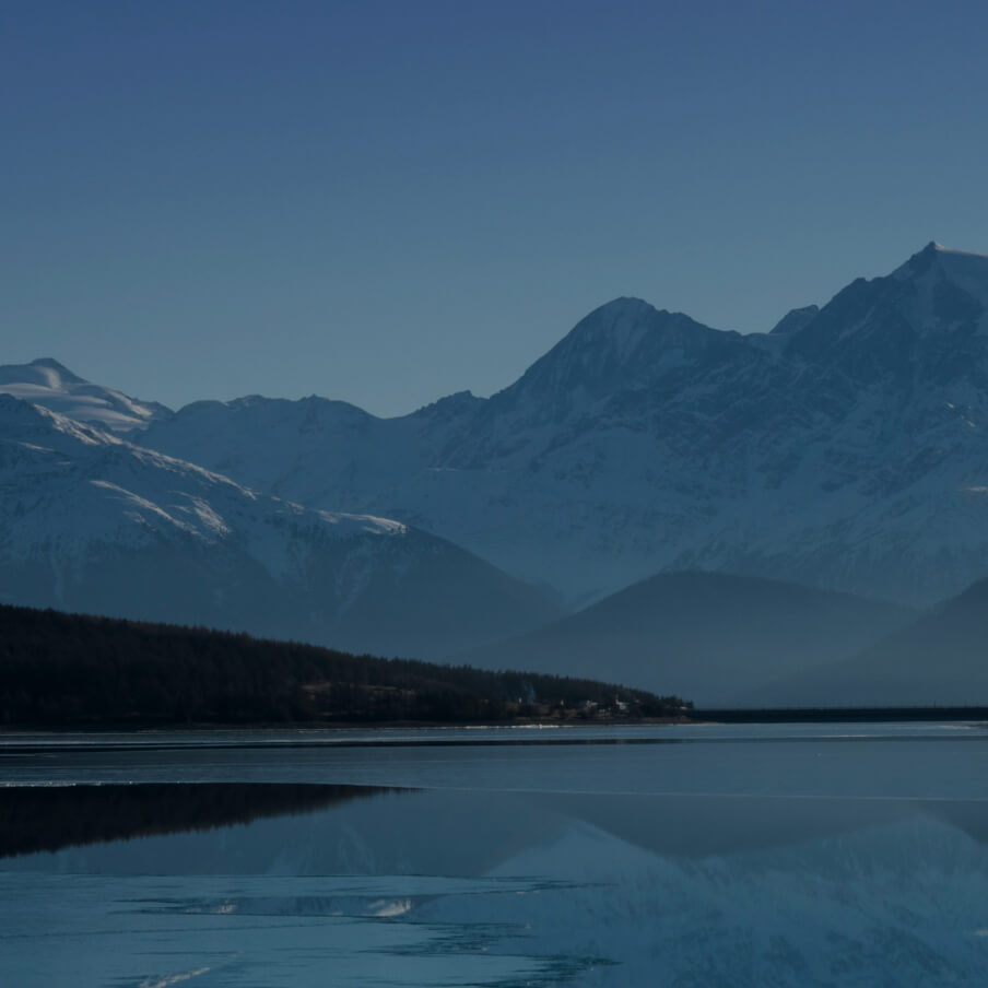 Calm lake reflecting a snow-covered mountain range under a clear blue sky.