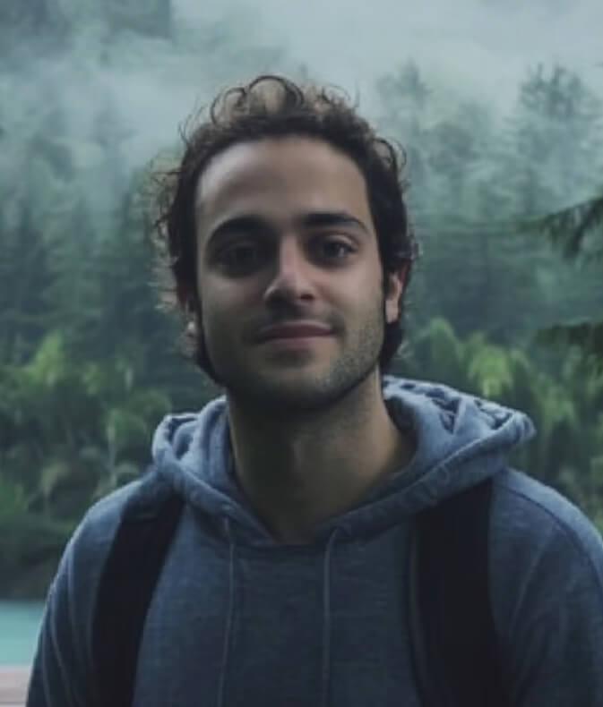 Young man with curly hair wearing a gray hoodie and backpack, standing outdoors with forest and mist in the background.