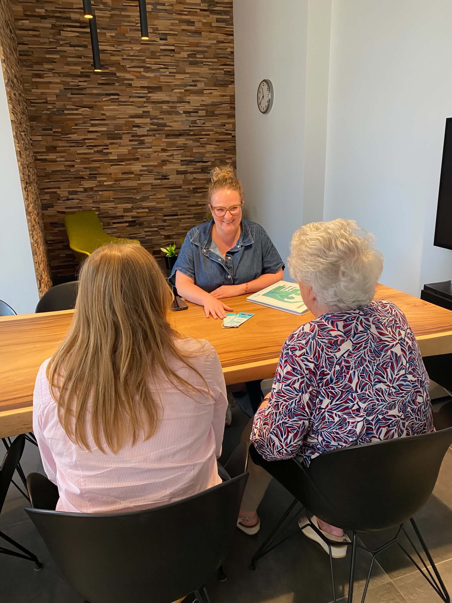 Three women seated around a wooden table in a modern room with wood-paneled wall; a younger woman wearing glasses and denim shirt smiles at two older women seated with their backs to the camera.
