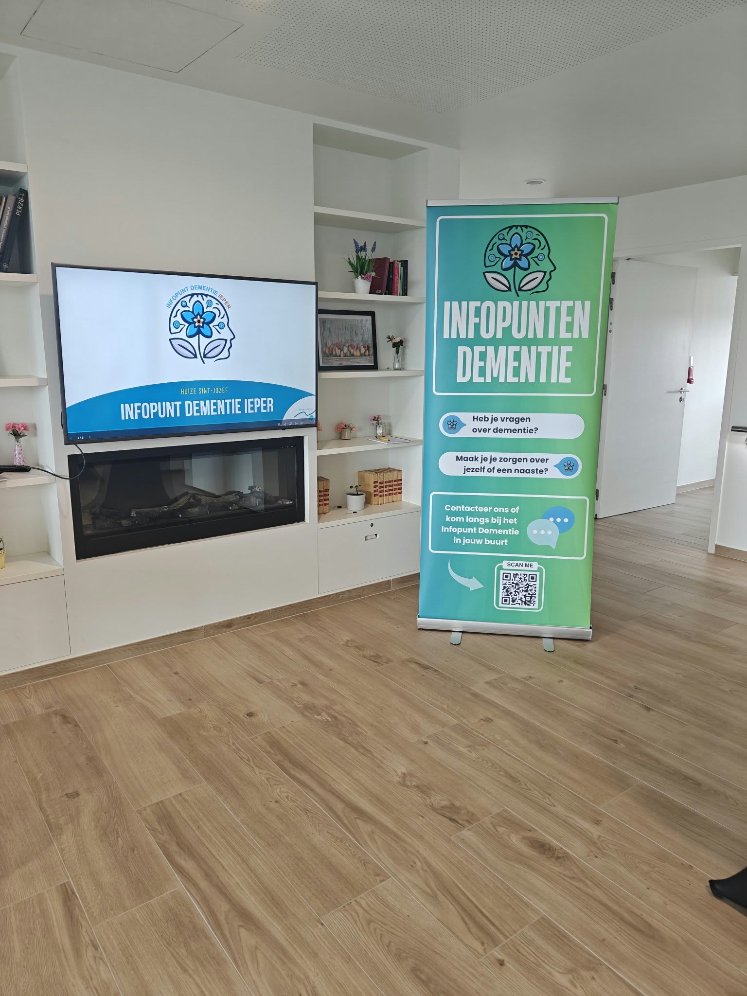Interior room with wooden flooring featuring a modern built-in fireplace beneath a screen displaying a logo and text 'Infopunt Dementie Ieper.' A blue-green standing banner beside the fireplace has the same logo and the text: Infopunten Dementie, two questions about dementia, and an invitation to contact or visit the local dementia information point with a QR code.