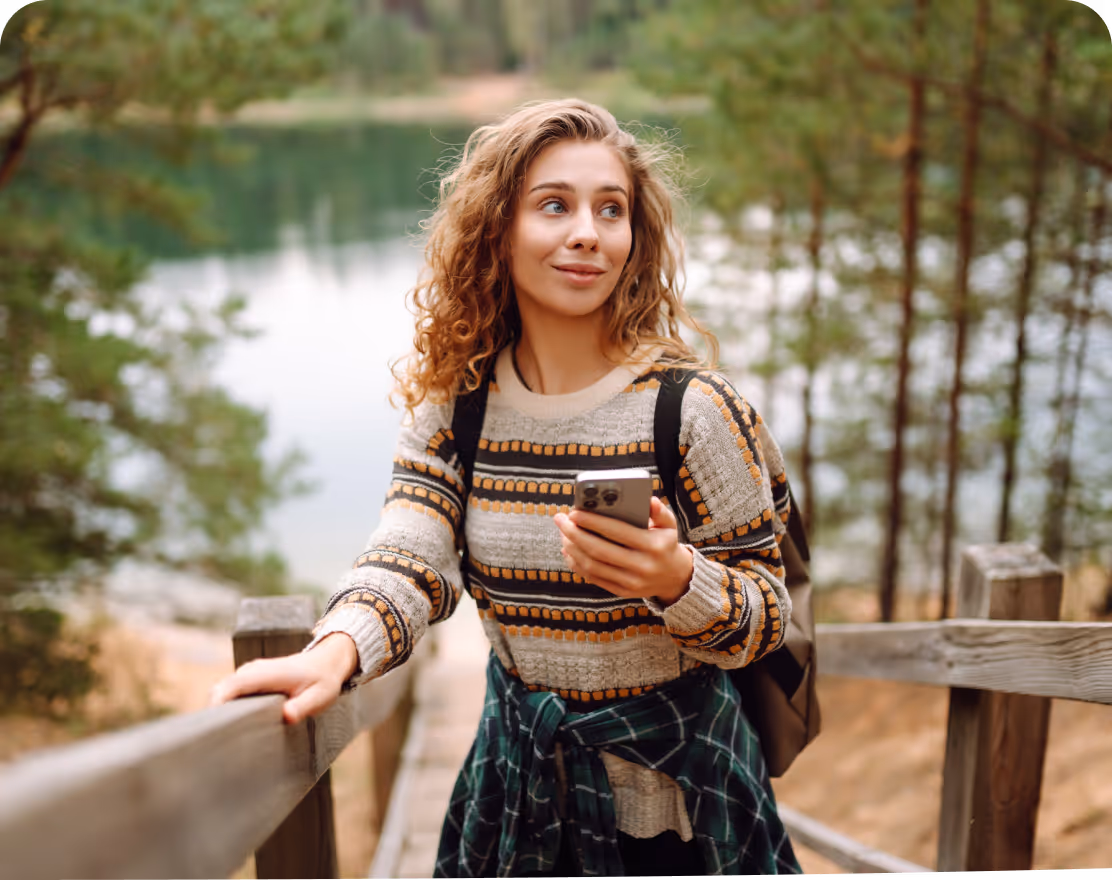 Jeune femme souriante en pull à motifs marchant sur un pont en bois en forêt, tenant un téléphone.