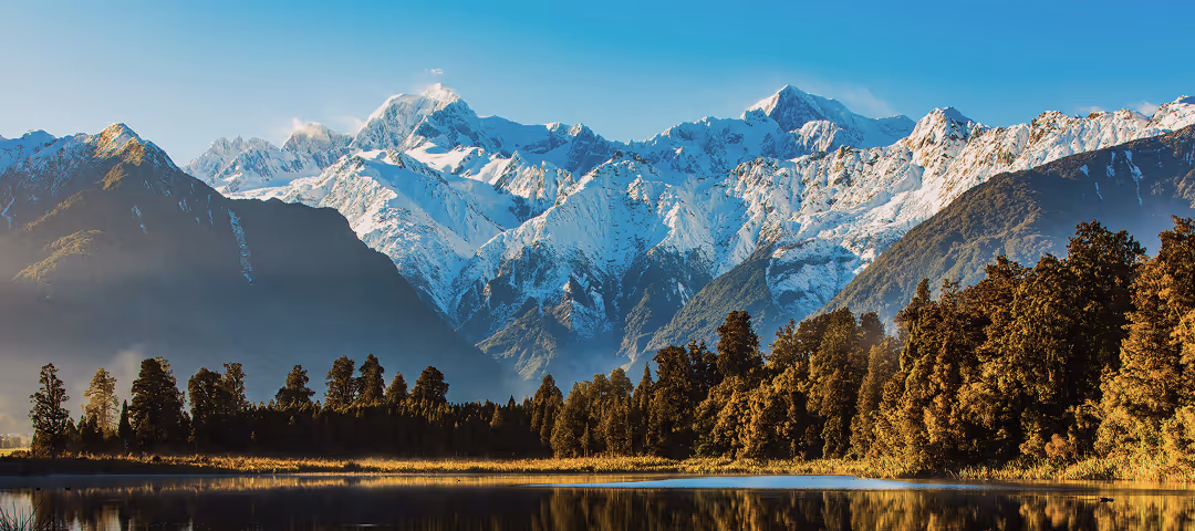 Montagnes enneigées majestueuses derrière une forêt dense avec un lac au premier plan reflétant le paysage sous un ciel clair.