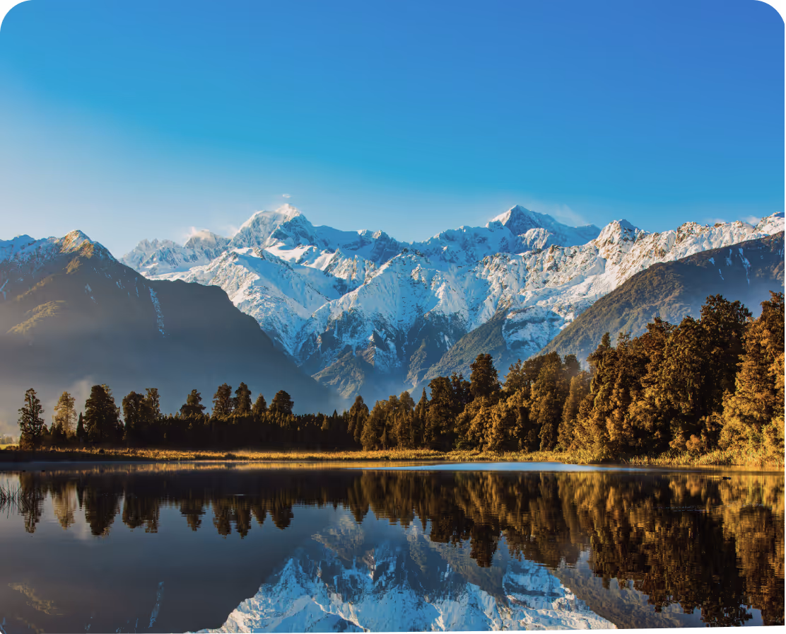 Montagnes enneigées avec forêt au premier plan et reflet dans un lac calme sous un ciel bleu clair.