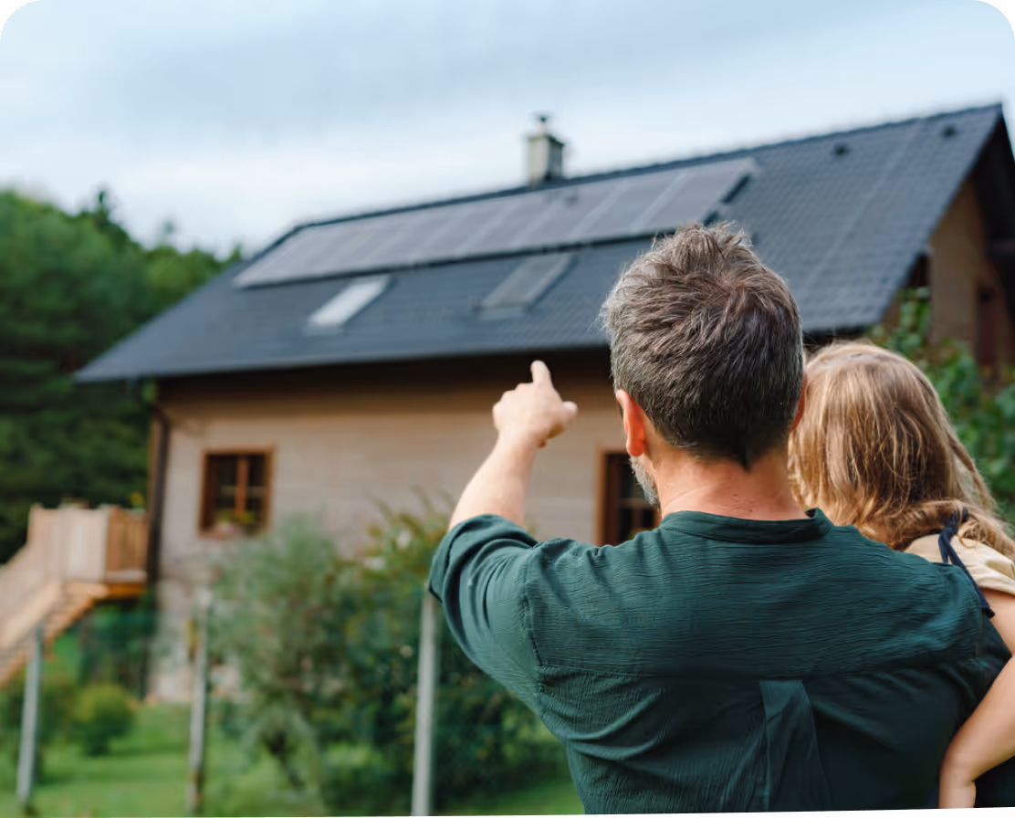 Un homme tenant une enfant pointe du doigt des panneaux solaires sur le toit d'une maison en bois.