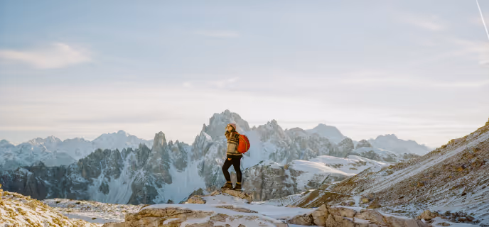 Une randonneuse debout sur une roche avec un sac à dos rouge, contemplant une chaîne de montagnes enneigées.