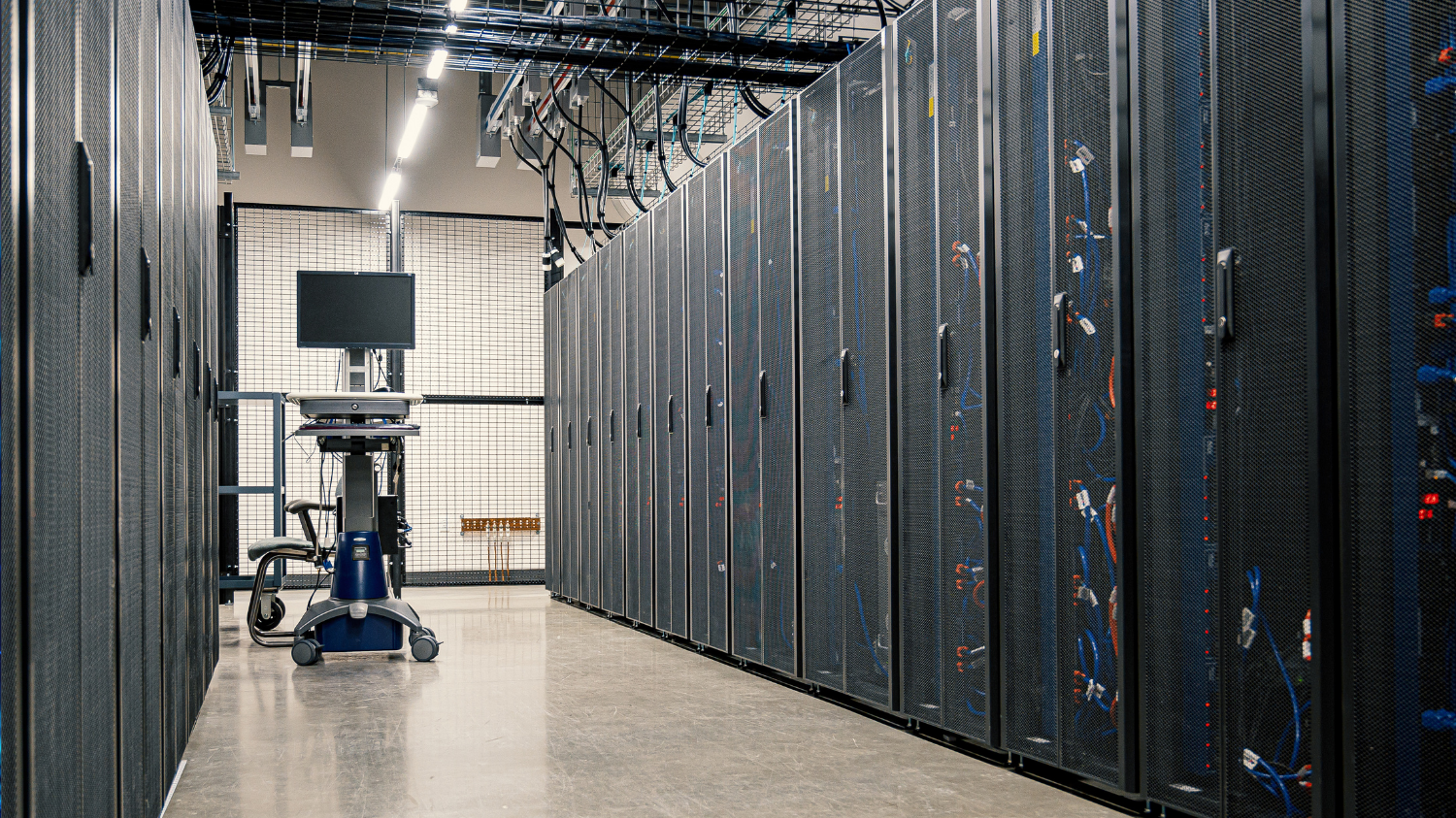 Allée d'un datacenter avec des baies de serveurs, un poste informatique mobile et des câbles réseau au plafond.