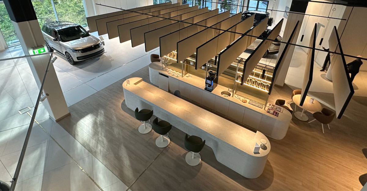 3D-illustrasjon av et moderne og lyst bilshowroom, med lyse treflater, sittegrupper og en rød bil utstilt i et eget område. Takhengte elementer skaper et luftig uttrykk i rommet.