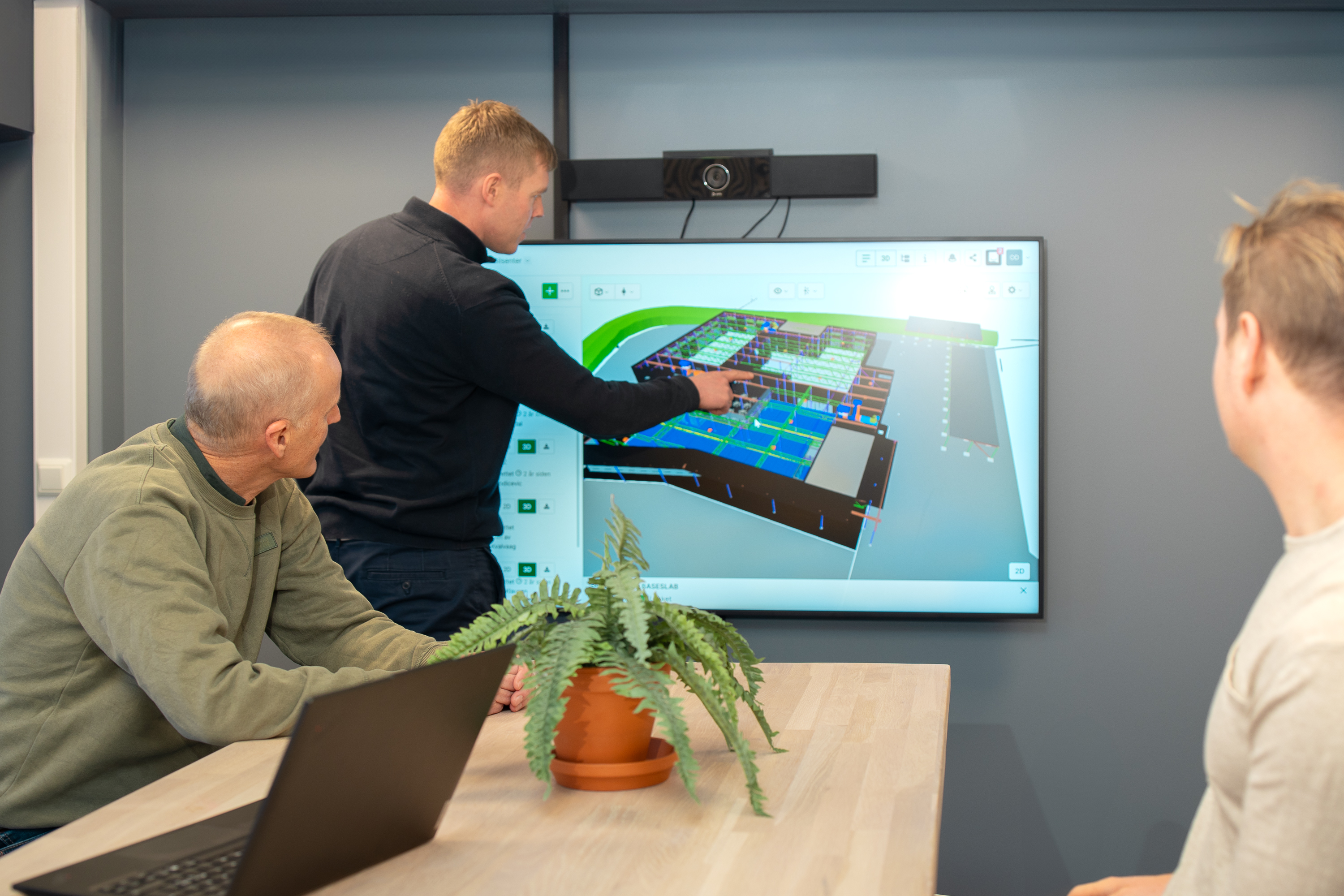 Tre Factor-ansatte i gule vernejakker og hjelmer står på en snødekt byggeplass med steinmasser i bakgrunnen. De diskuterer prosjektet og konsulterer papirer eller en PC.