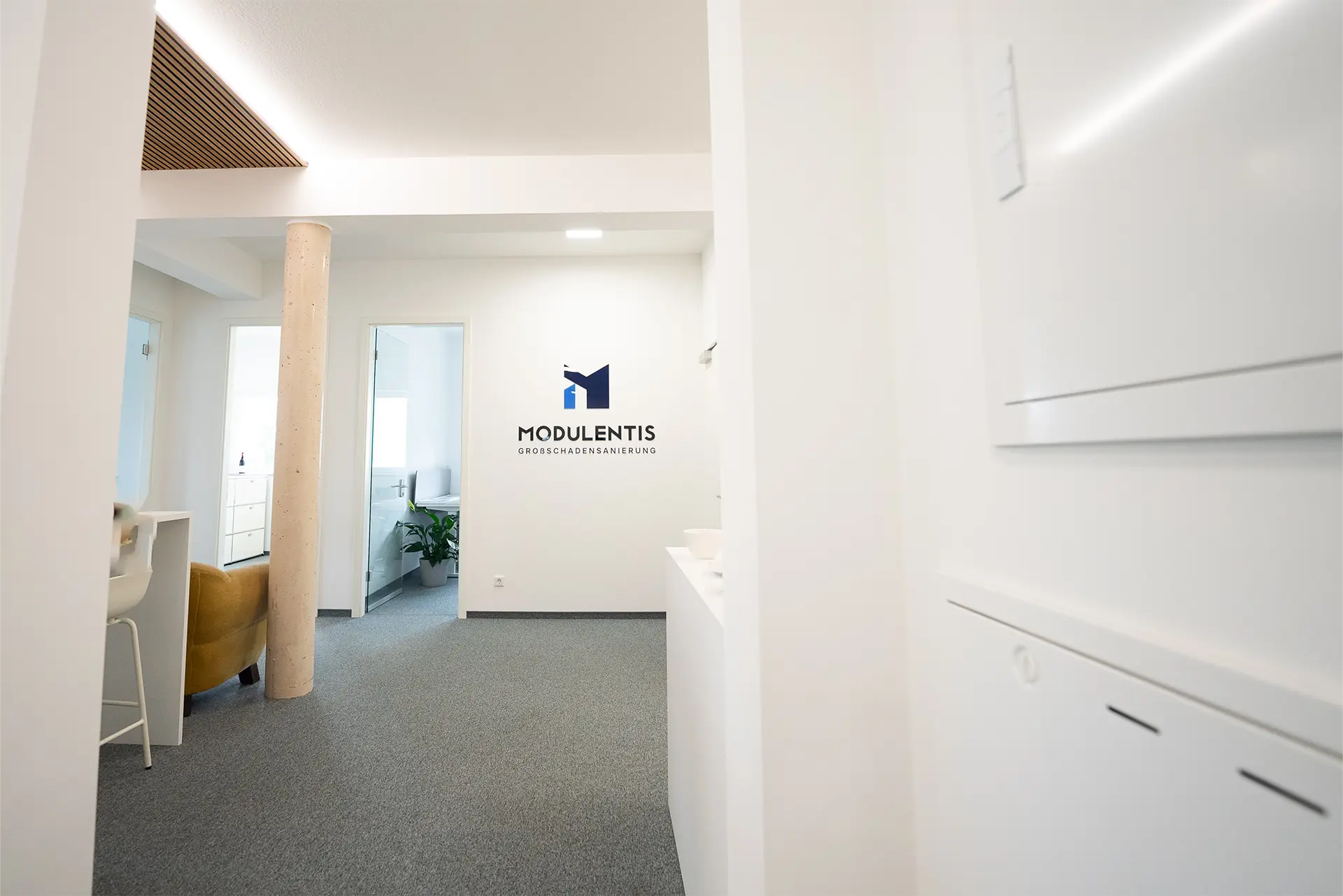 Blick in ein modernes Büro mit grauem Teppichboden. Links steht ein weißer Schreibtisch mit einem Stuhl. An der Wand ist das Logo 'MODULENTIS' zu sehen.