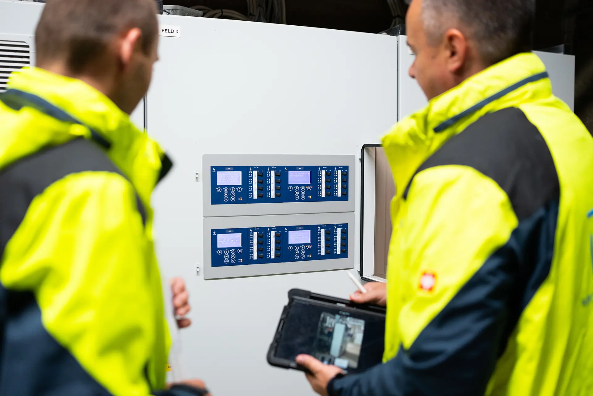 Zwei Personen in gelben Sicherheitsjacken stehen vor einem Bedienfeld mit mehreren Tasten und Displays. Eine Person hält ein Tablet in der Hand.