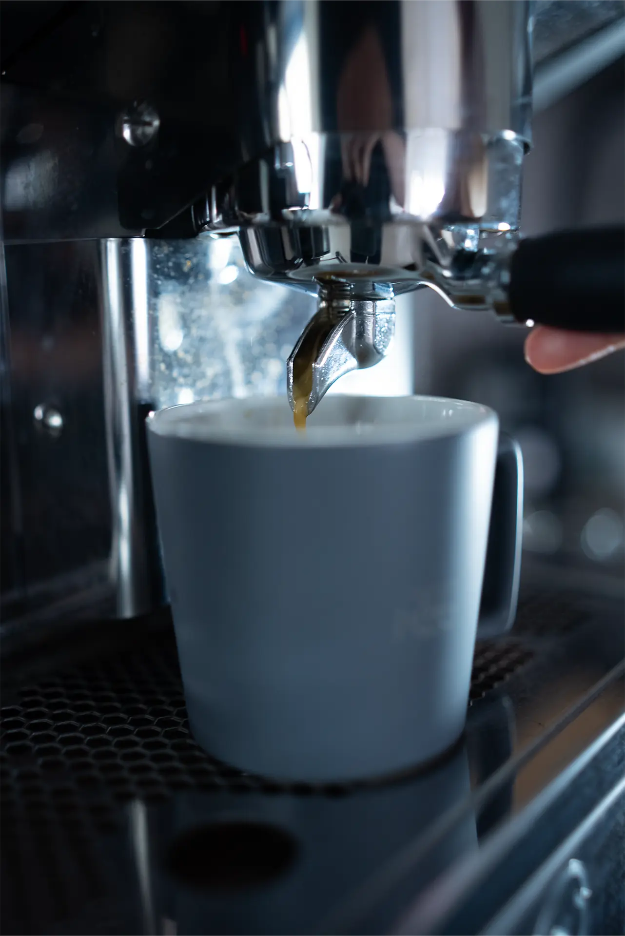 Eine Hand hält einen schwarzen Griff eines Siebträgers einer Kaffeemaschine, während ein Espresso in eine graue Tasse fließt. Die Maschine hat eine glänzende Metalloberfläche.