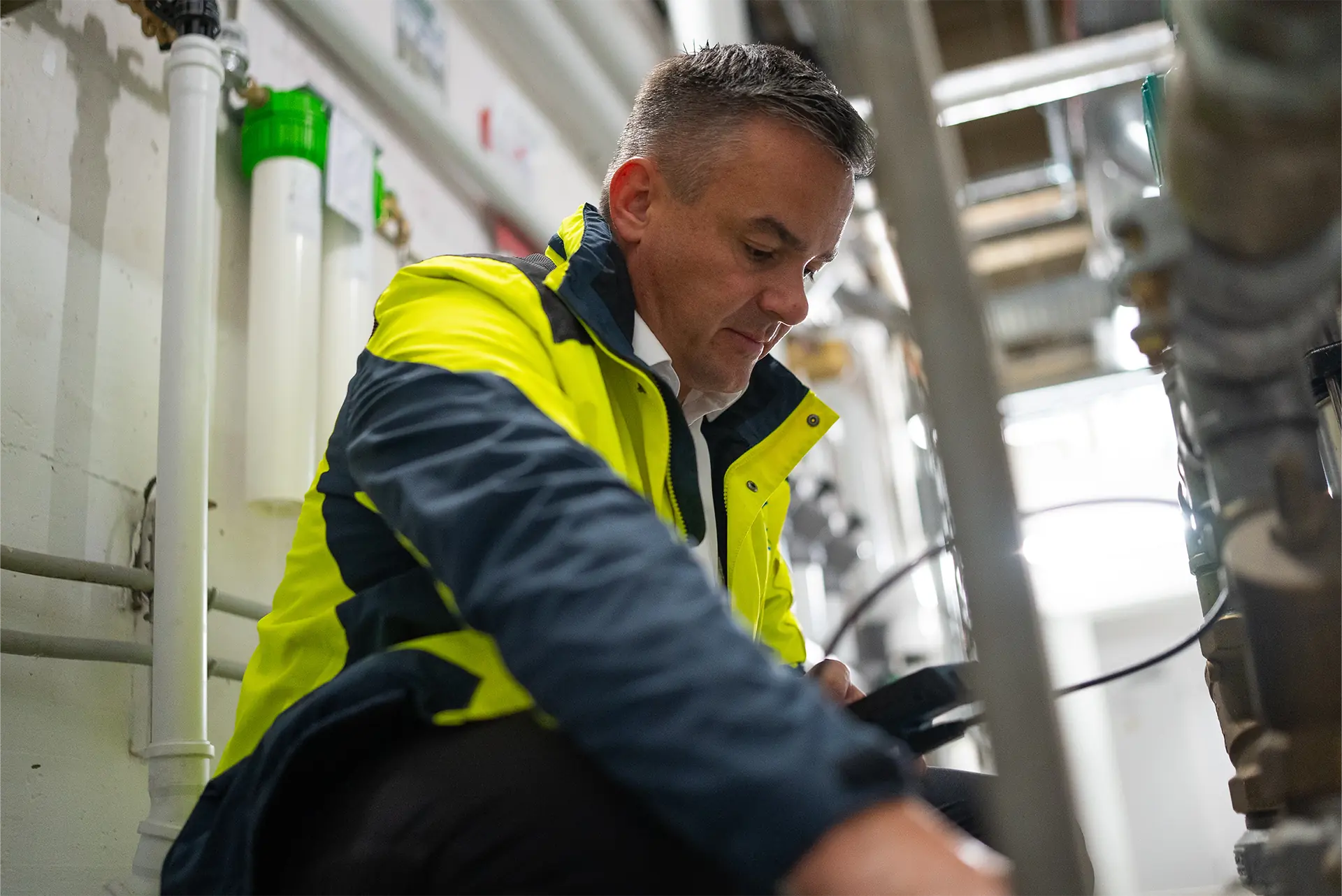 Eine Person in einer gelben Sicherheitsjacke sitzt auf dem Boden und arbeitet an Rohrleitungen in einer Industrieumgebung.