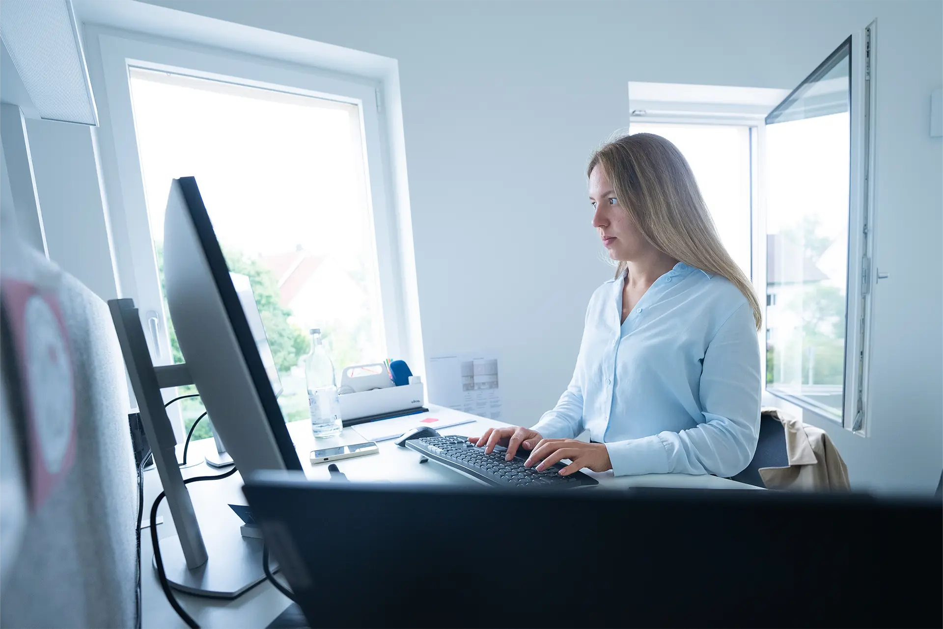 Eine Person sitzt an einem Schreibtisch und tippt auf einer Tastatur. Auf dem Tisch stehen zwei Computerbildschirme und eine Wasserflasche.
