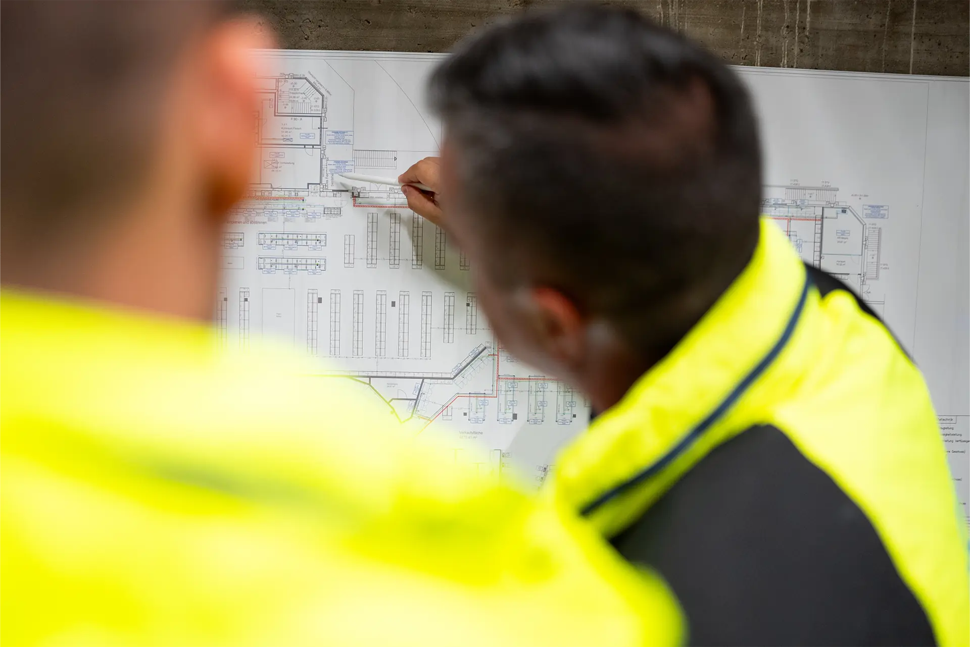 Zwei Personen in gelben Sicherheitsjacken betrachten einen Bauplan. Eine Person zeigt mit einem Stift auf einen bestimmten Bereich des Plans.