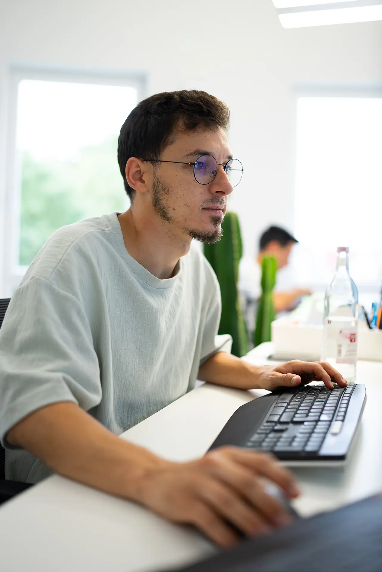 Ein Mann mit Brille sitzt an einem Schreibtisch und arbeitet an einer Tastatur. Im Hintergrund sind weitere Personen und Pflanzen sichtbar.