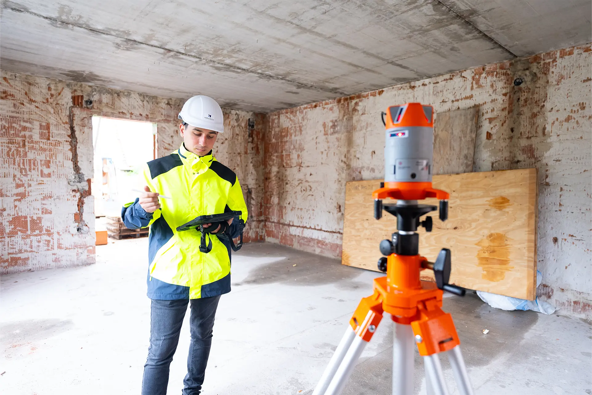 Person in leuchtend gelber Sicherheitsjacke und weißem Helm steht neben einem orangefarbenen Nivelliergerät auf einem dreibeinigen Stativ in einem Rohbau und prüft Daten auf einem Tablet.