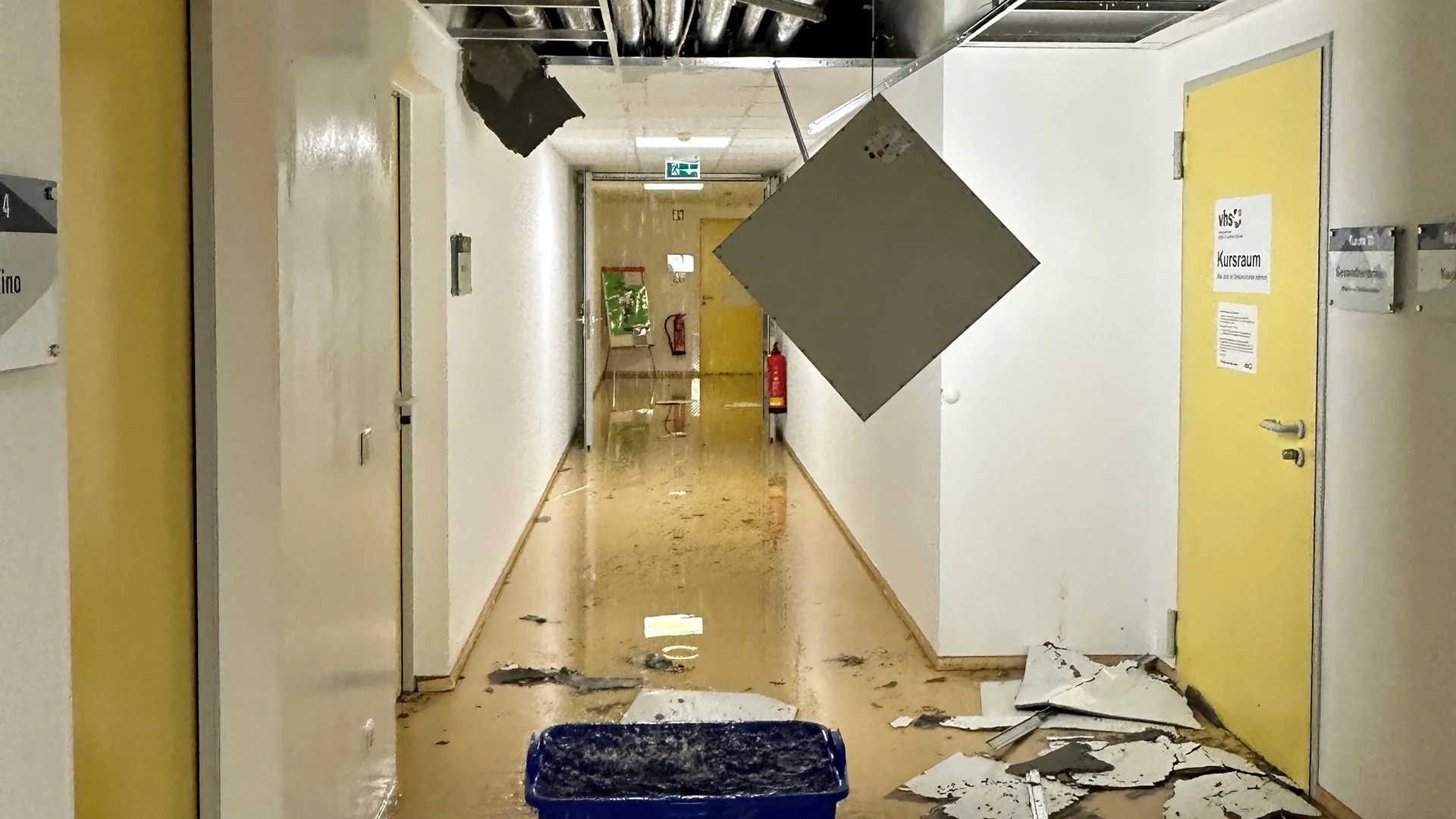 Flur nach Elementarschaden mit beschädigter Decke, herabhängender Deckenplatte und Schutt auf dem Boden, gelbe Tür mit Schild 'Kursraum'