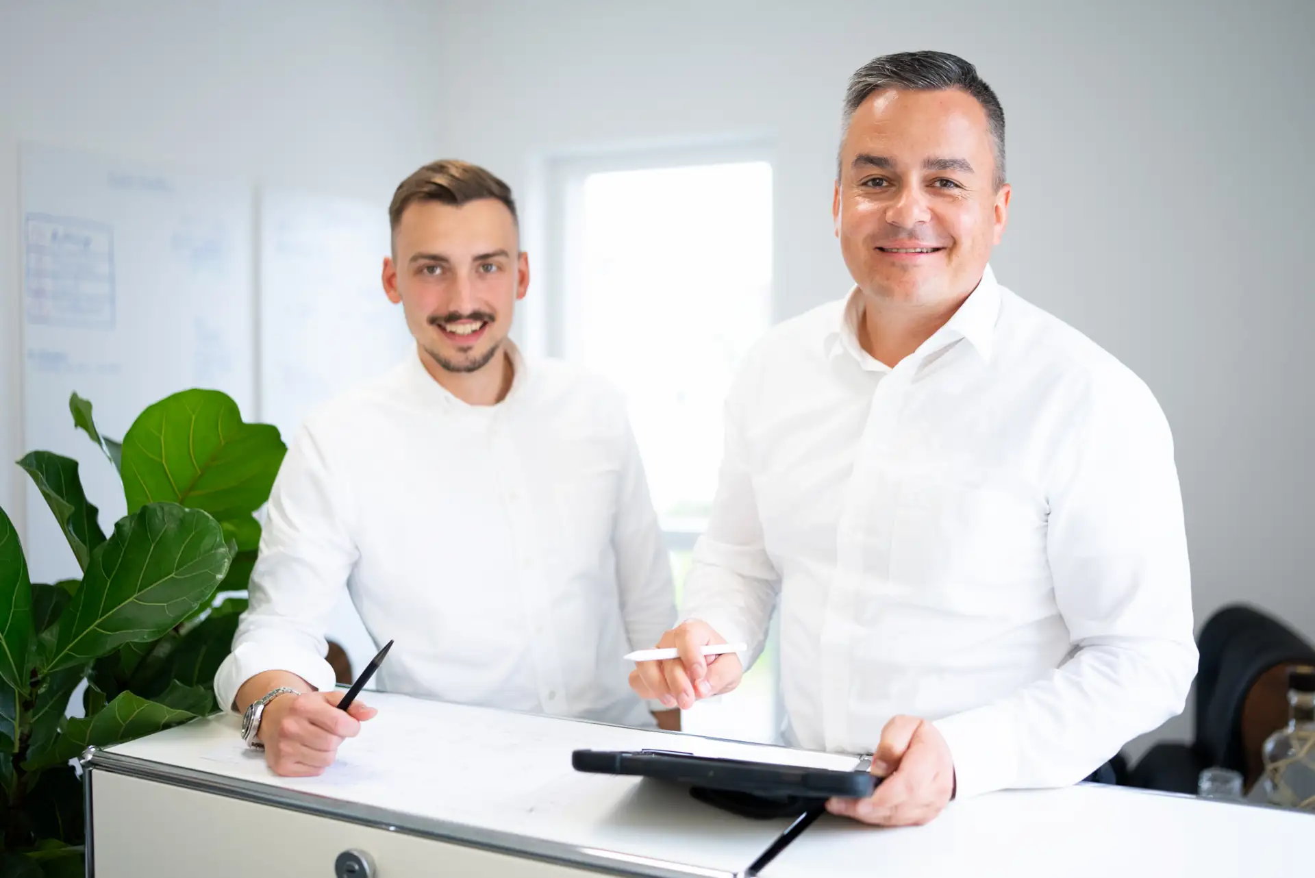 Sebastian Erlbeck und Matthias Röck in weißen Hemden stehen an einem Tisch mit Unterlagen und Stiften in einem hellen Büro