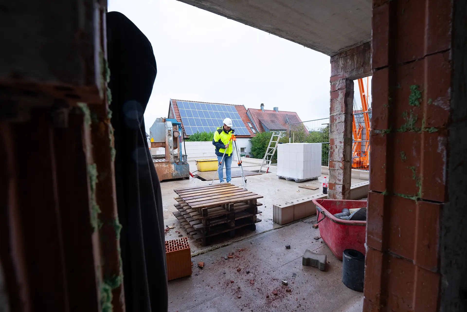 Bauarbeiter mit Helm und Warnweste steht auf einer Baustelle vor einem Haus mit Solarpaneelen auf dem Dach