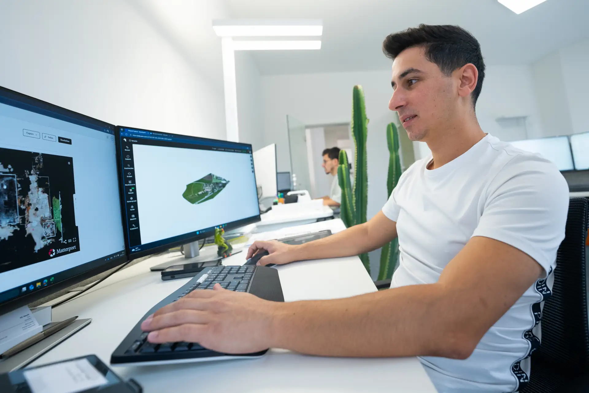 Junger Mann sitzt an Schreibtisch mit zwei Monitoren und arbeitet an 3D-Scans eines Schadenfalls in hellem Büro