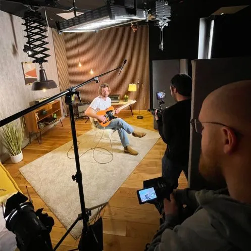 Ein Mann sitzt auf einem Stuhl und spielt in einem Aufnahmestudio E-Gitarre, während zwei Kameramänner ihn filmen.