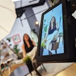 Auf dem Kamerabildschirm ist eine Frau zu sehen, die während eines Videodrehs in einem hellen Raum auf einem Hocker sitzt.