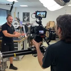 Bildschirm einer Videokamera, auf dem ein Mann mit Duttfrisur in einem hell erleuchteten Büro mit Computertischen zu sehen ist.