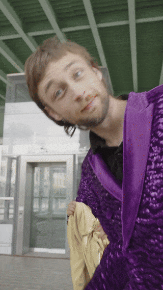 Ein Mann in einer glänzenden lila Jacke beugt sich vor einem Gebäude mit Glastüren zur Kamera.