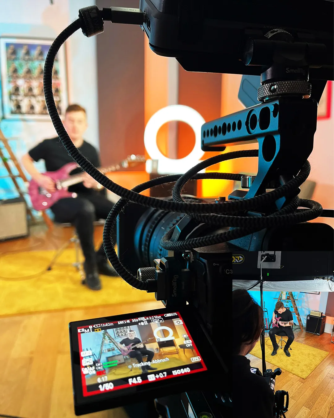 Die Videokamera ist auf einen Mann gerichtet, der in einem Studio mit warmem Licht und einem Ringlicht im Hintergrund E-Gitarre spielt.