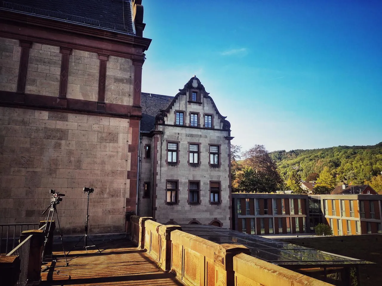 Historisches Steingebäude mit großen Fenstern, Außenkamera und Beleuchtung auf einem Balkon, im Hintergrund moderne Gebäude mit grünen Bäumen unter klarem blauen Himmel.