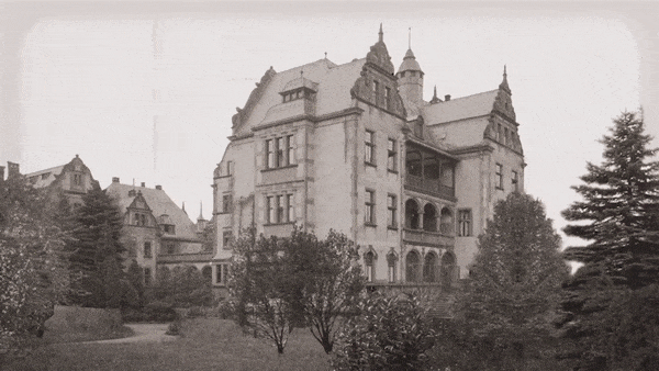 Schwarz-Weiß-Video einer großen historischen Villa mit aufwendiger Architektur, umgeben von Bäumen und einem Gartenweg.