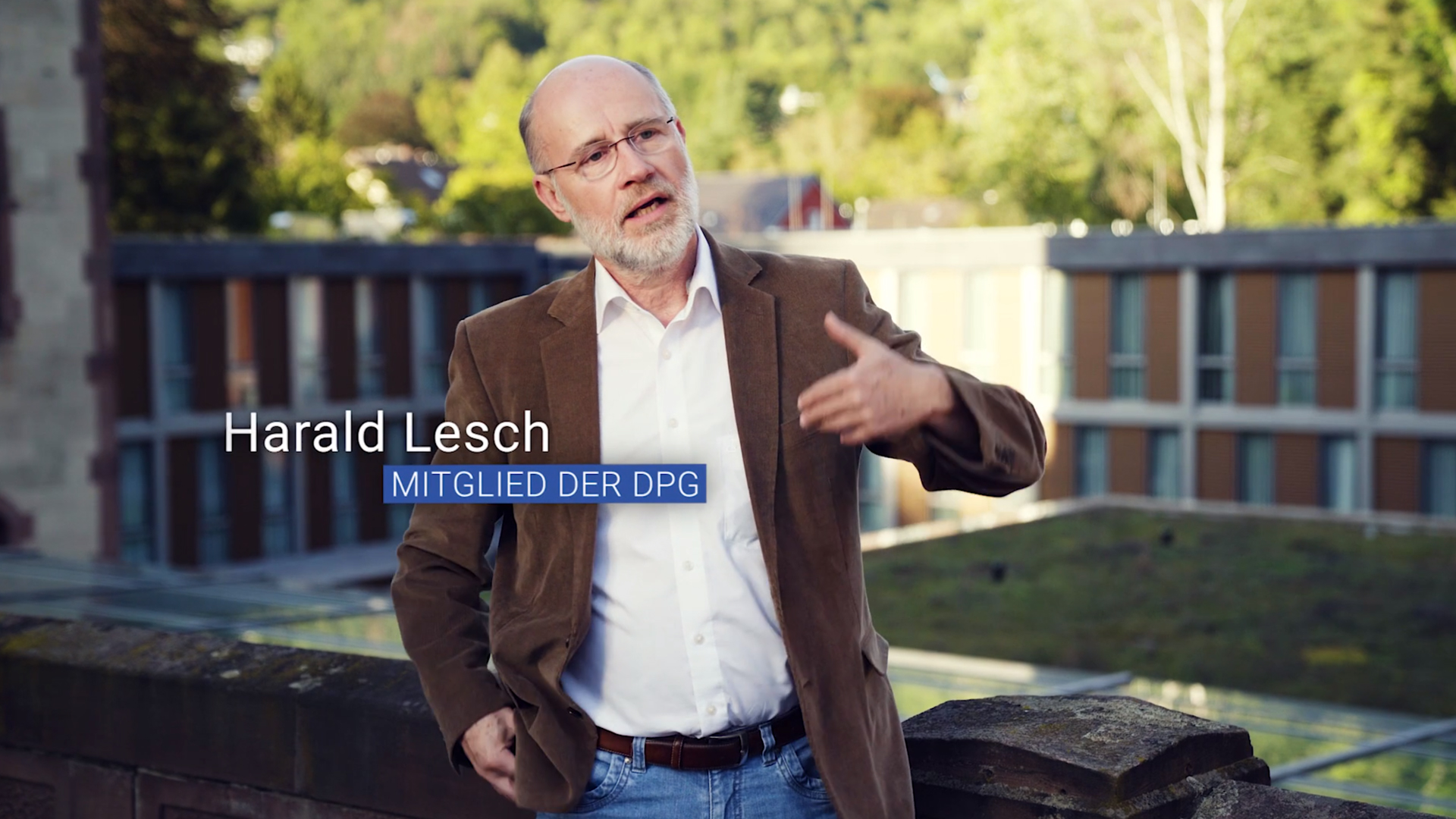Ein Mann mittleren Alters mit Brille und Bart spricht im Freien; er trägt eine braune Jacke und ein weißes Hemd.