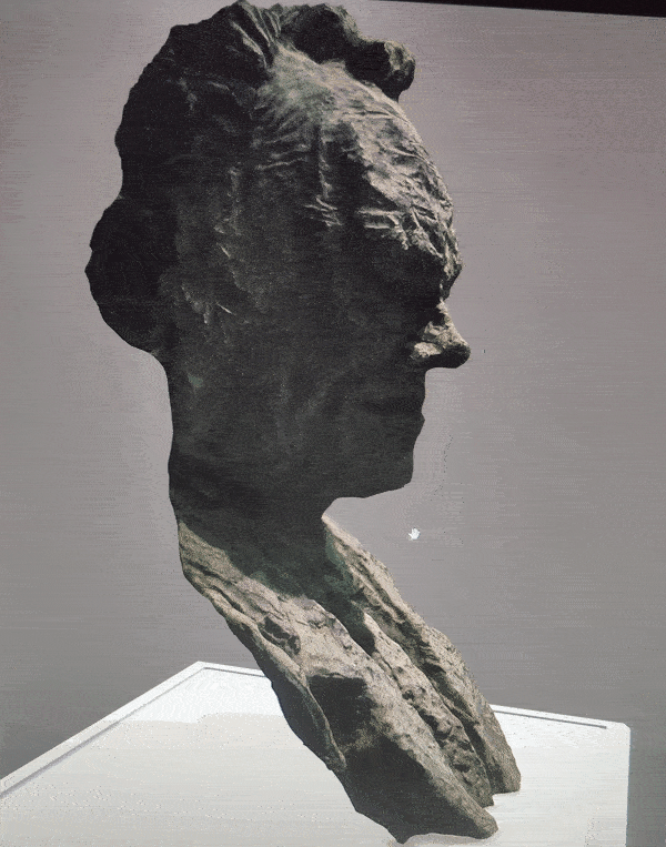 Drehbare Bronzebüste eines Mannes mit strukturiertem Haar und detaillierten Gesichtszügen auf einem weißen Sockel vor grauem Hintergrund.