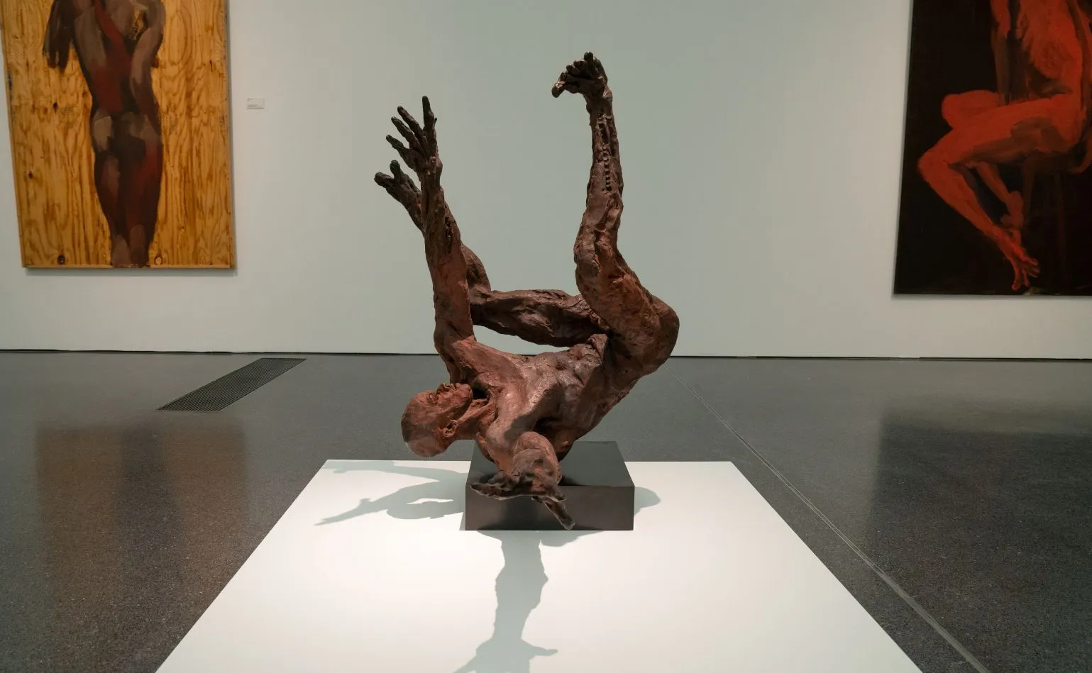 Abstrakte, dunkelbronzene Skulptur einer verrenkten menschlichen Figur auf einem schwarzen Sockel in einer Galerie mit Gemälden an den Wänden.