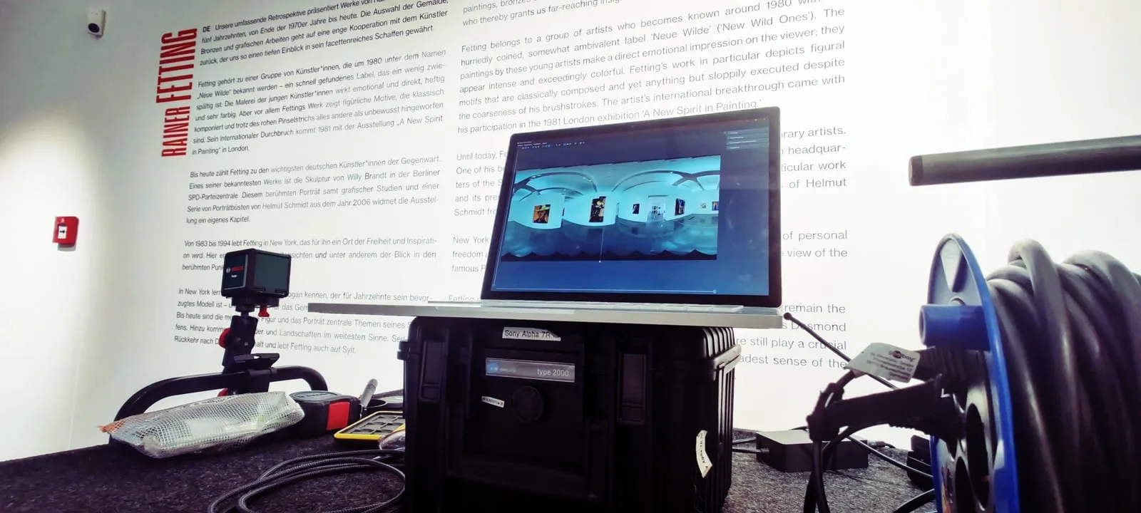 Auf einem schwarzen Hartschalenkoffer steht ein Laptop mit Fotobearbeitungssoftware, daneben eine GoPro-Kamera an einem Griff und eine große Kabeltrommel. Das Ganze steht vor einer weißen Wand, die mit deutschen und englischen Texten über die Kunst von Rainer Fetting bedeckt ist.