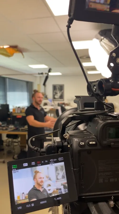 Bildschirm einer Videokamera, auf dem ein Mann mit Duttfrisur in einem hell erleuchteten Büro mit Computertischen zu sehen ist.