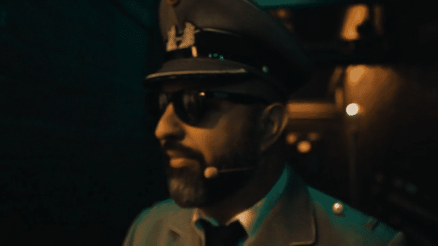 Mann mit Militärmütze, Sonnenbrille und Mikrofon-Headset vor einer Bühne.