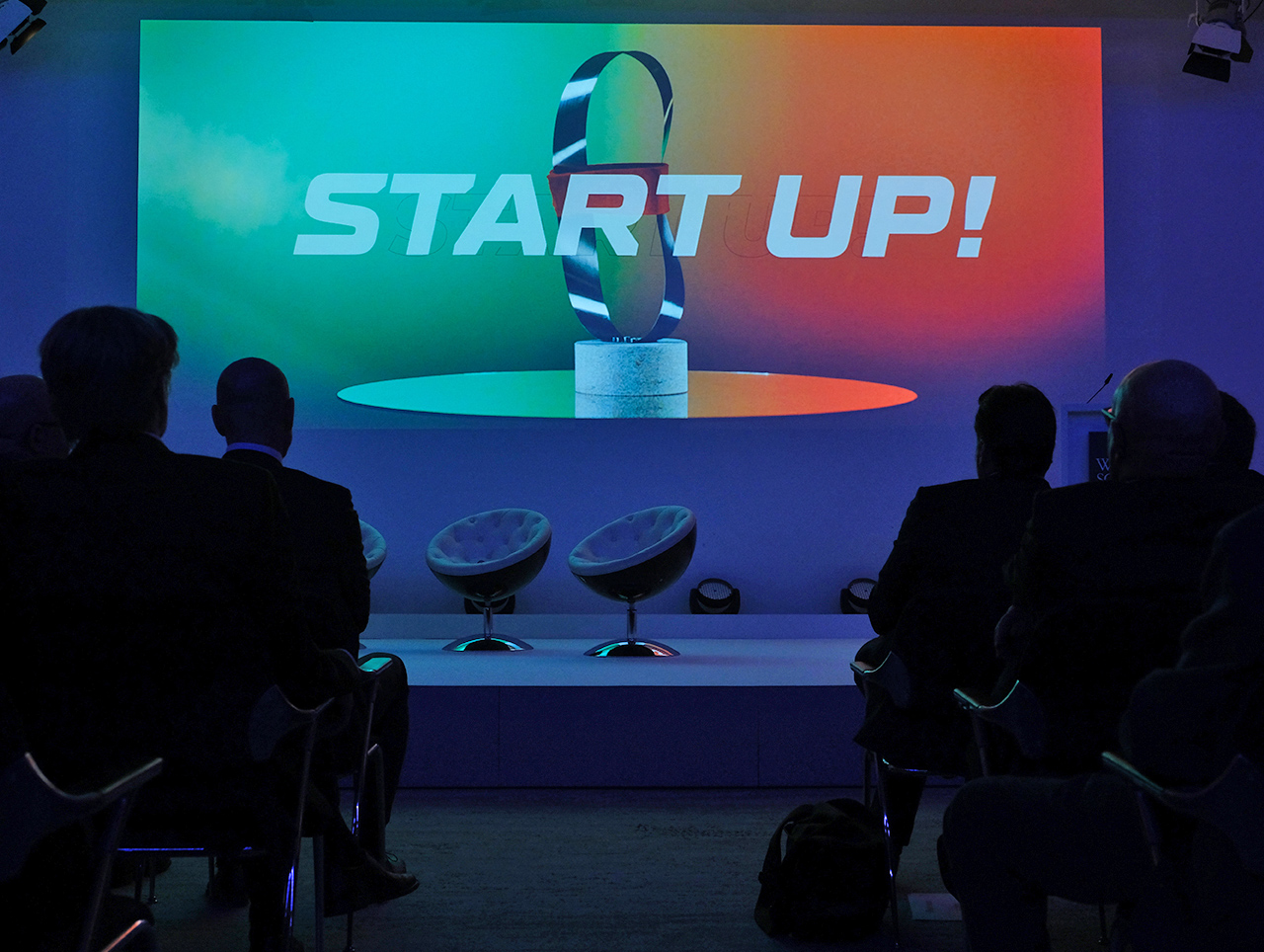 Zuschauer sitzen in einem schwach beleuchteten Raum und betrachten einen farbenfrohen Bildschirm, auf dem eine metallische Bandskulptur und der Text „START UP!“ zu sehen sind.