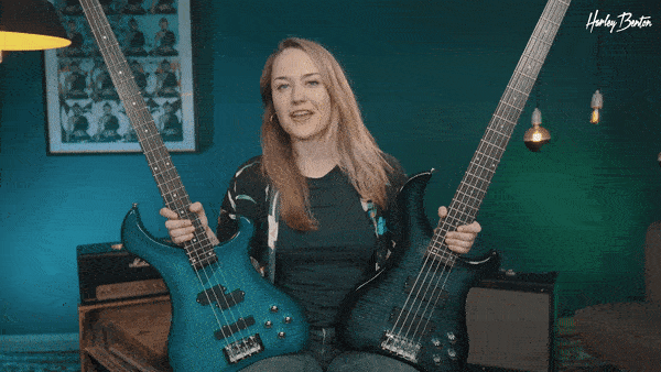 Frau sitzt drinnen, hält zwei elektrische Bassgitarren in der Hand und präsentiert sie, lächelt und spricht in die Kamera.