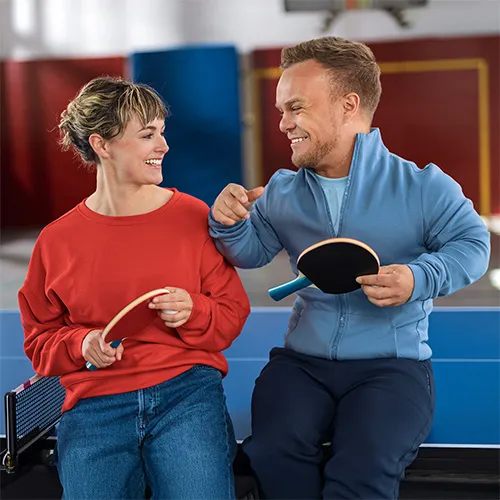 Ein lächelnder Mann und eine lächelnde Frau sitzen an einer Tischtennisplatte und unterhalten sich angeregt.