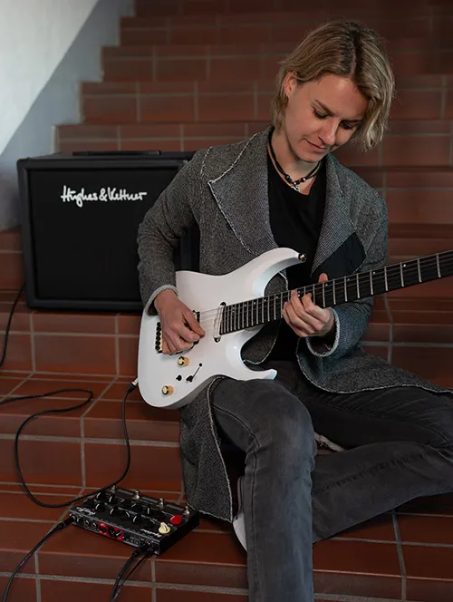 Eine Frau sitzt auf einer Treppe und spielt eine weiße E-Gitarre; im Hintergrund ist ein Hughes & Kettner-Verstärker zu sehen.