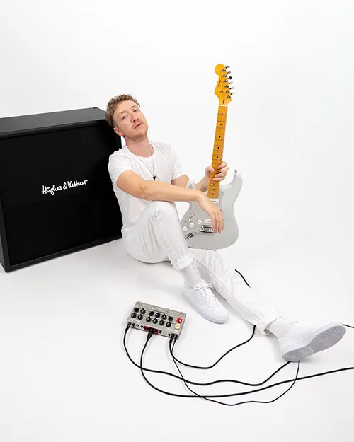 Ein Mann, ganz in Weiß gekleidet, sitzt auf dem Boden und hält eine weiße E-Gitarre neben einem Hughes & Kettner Verstärker und einem Pedalboard mit angeschlossenen Kabeln.
