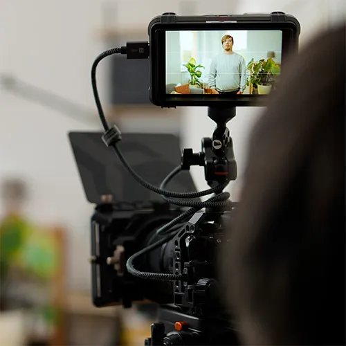 Kameraaufbau zur Aufnahme einer Person, die in einem Innenraum steht; im Hintergrund sind Pflanzen zu sehen, die auf dem Monitor der Kamera angezeigt werden.